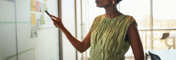 Focused businesswoman presents ideas on a whiteboard covered in sticky notes. She points to a note, amidst geometric lines on the board, bathed in natural office light.
